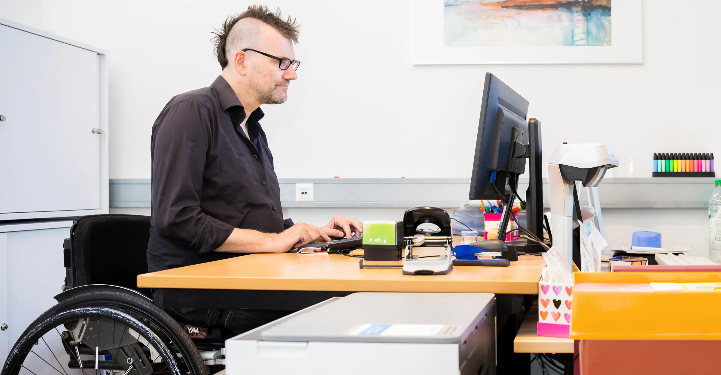 Bürosituation:  Herr Freels sitzt am Schreibtisch und schreibt am Computer. Er trägt ein schwarzes Hemd.