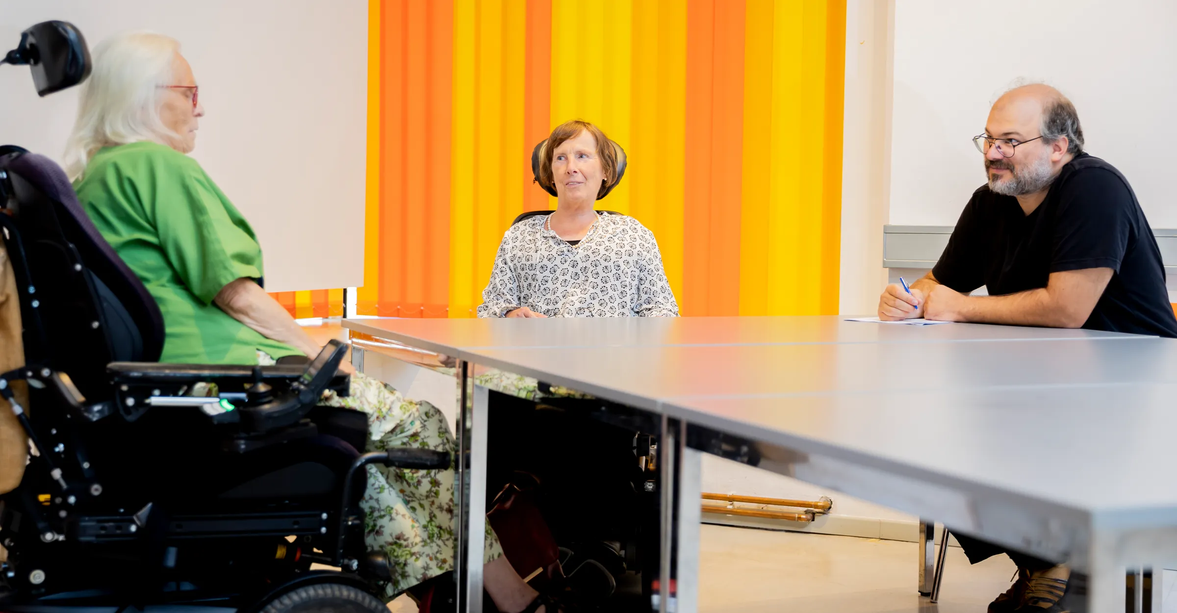 Zu sehen ist eine Beratungssituation: im Hintergrund sieht man orangefarbene Lamellenvorhänge. Es gibt einen länglichen, grauen Tisch. Auf der linken Seite sitzt die Klientin, in der Bildmitte sitzt Frau Stenger, auf der rechten Seite vom Tisch sitzt Frau Stengers Assistenz.