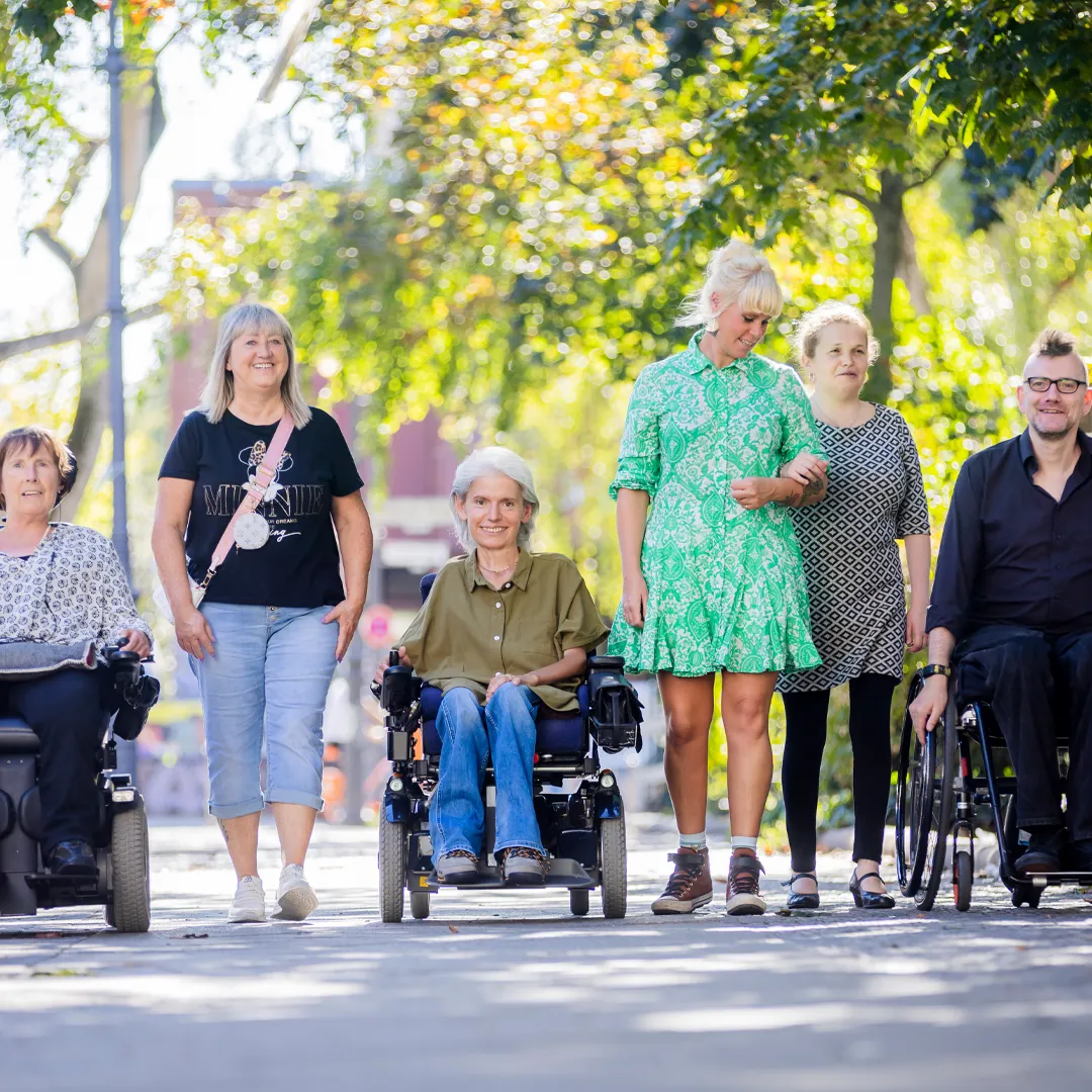 Teamfoto, 6 Personen laufen in sommerlicher Kleidung, lächelnd auf den Betrachter zu. Die Sonne scheint von hinten durch die Bäume. Von links nach rechts: Frau Stenger, eine Assistentin, Frau Peric, eine Assistentin, Frau Merslikin und Herr Freels. Frau Peric und Frau Stenger fahren im Elektrorollstuhl, Frau Merslikin hat sich bei der Assistentin untergehakt, Herr Freels fährt einen Aktivrollstuhl.