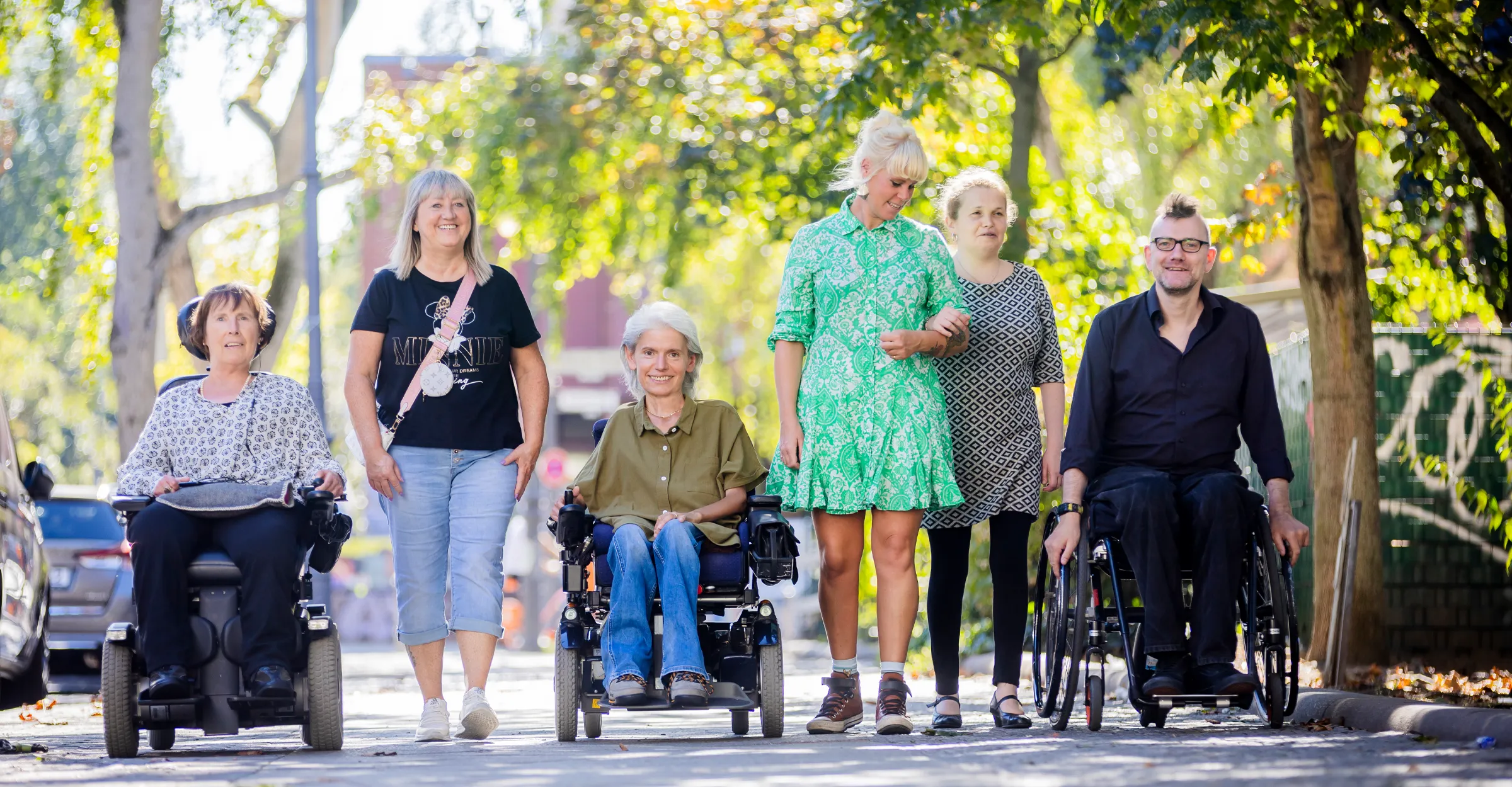 Teamfoto, 6 Personen laufen in sommerlicher Kleidung, lächelnd auf den Betrachter zu. Die Sonne scheint von hinten durch die Bäume. Von links nach rechts: Frau Stenger, eine Assistentin, Frau Peric, eine Assistentin, Frau Merslikin und Herr Freels. Frau Peric und Frau Stenger fahren im Elektrorollstuhl, Frau Merslikin hat sich bei der Assistentin untergehakt, Herr Freels fährt einen Aktivrollstuhl.