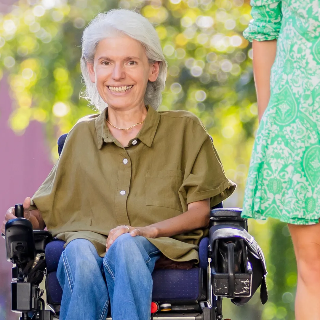 Auf der linken Bildhälfte sieht man Frau Peric in die Kamera lächeln. Sie trägt eine olivfarbene Bluse. Rechts von ihr im Anschnitt eine Assistentin, die ein hellgrünes Kleid trägt. Unscharf im Hintergrund sieht man Bäume.