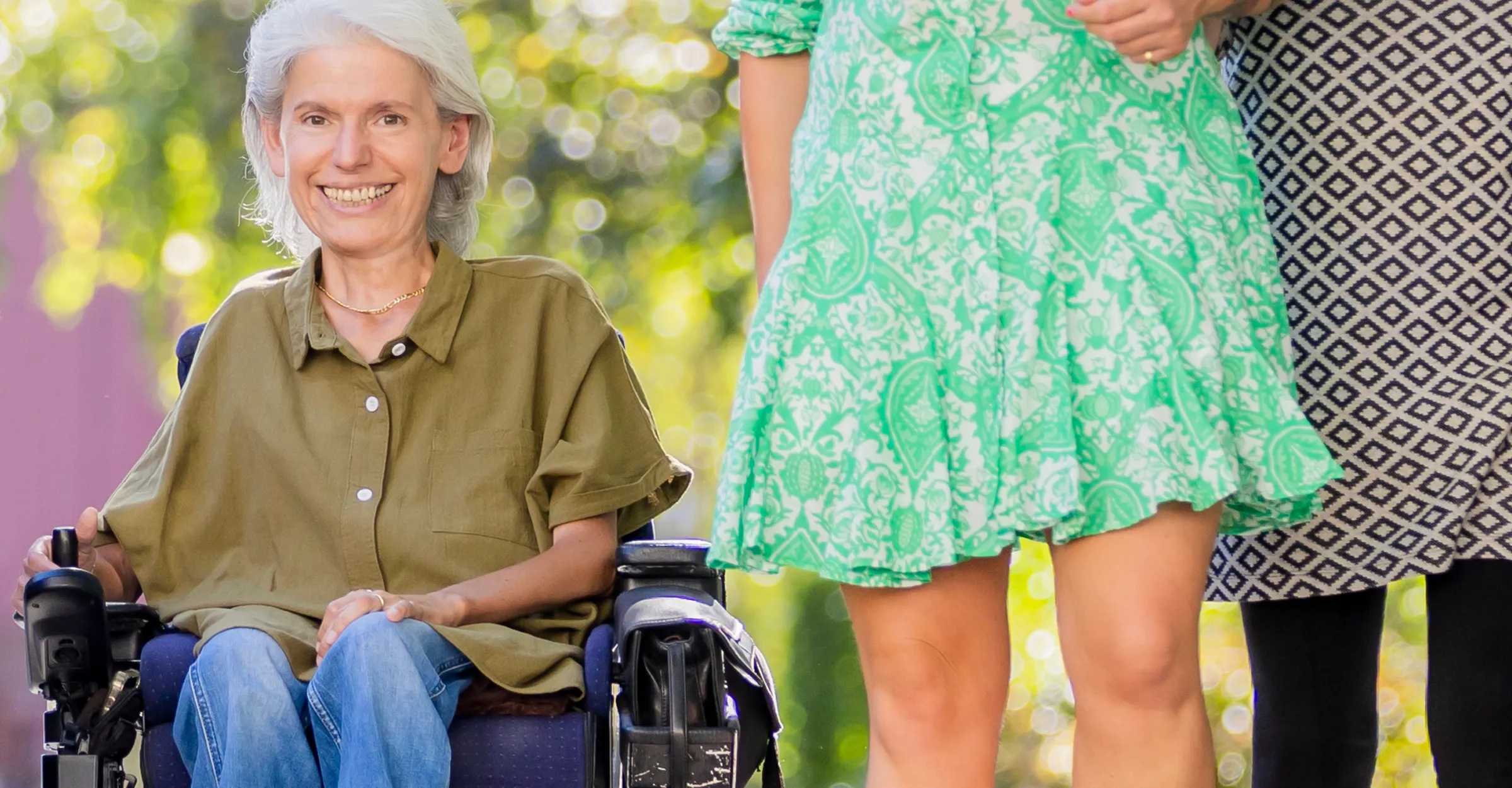 Auf der linken Bildhälfte sieht man Frau Peric in die Kamera lächeln. Sie trägt eine olivfarbene Bluse. Rechts von ihr im Anschnitt eine Assistentin, die ein hellgrünes Kleid trägt. Unscharf im Hintergrund sieht man Bäume.
