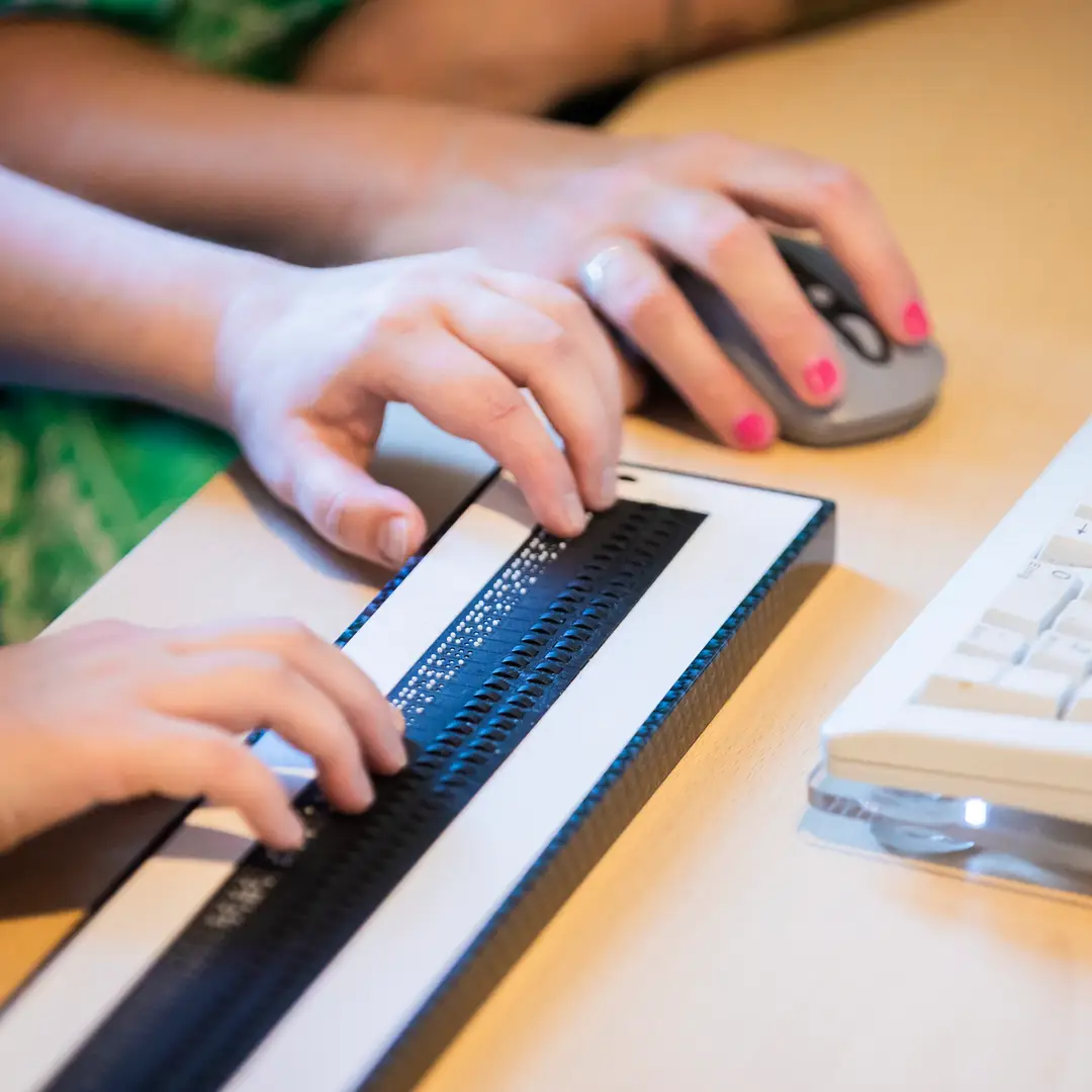 Nahaufnahme Schreibtisch: im Anschnitt ist eine Tastatur zu sehen, eine Braillezeile und eine Maus. Zwei Hände lesen auf der Braillezeile, eine Hand bedient die Maus.