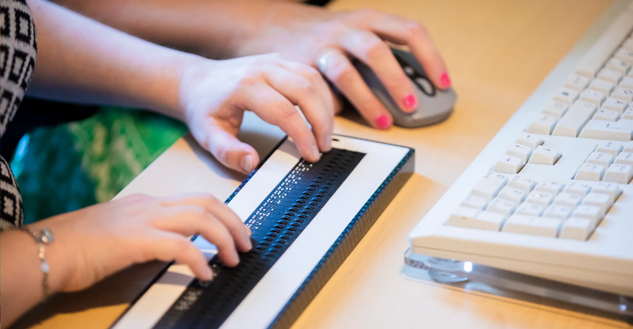 Nahaufnahme Schreibtisch: im Anschnitt ist eine Tastatur zu sehen, eine Braillezeile und eine Maus. Zwei Hände lesen auf der Braillezeile, eine Hand bedient die Maus.