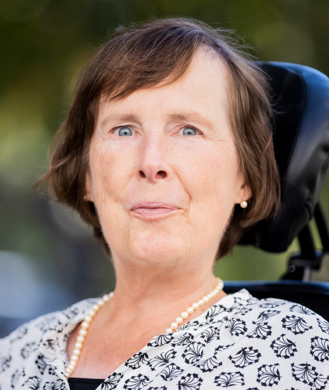 Frau Stenger hat kinnlanges, braunes Haar mit einem Pony, der zur Seite gekämmt ist. Sie hat hellblaue Augen, trägt weiße Perlenohrringe und eine weiße Perlenkette. Sie trägt eine weiße Bluse, die mit dunkelblauen Blumen gemustert ist. Sie lächelt frontal in die Kamera. Im Hintergrund sieht man unscharf grüne Pflanzen.