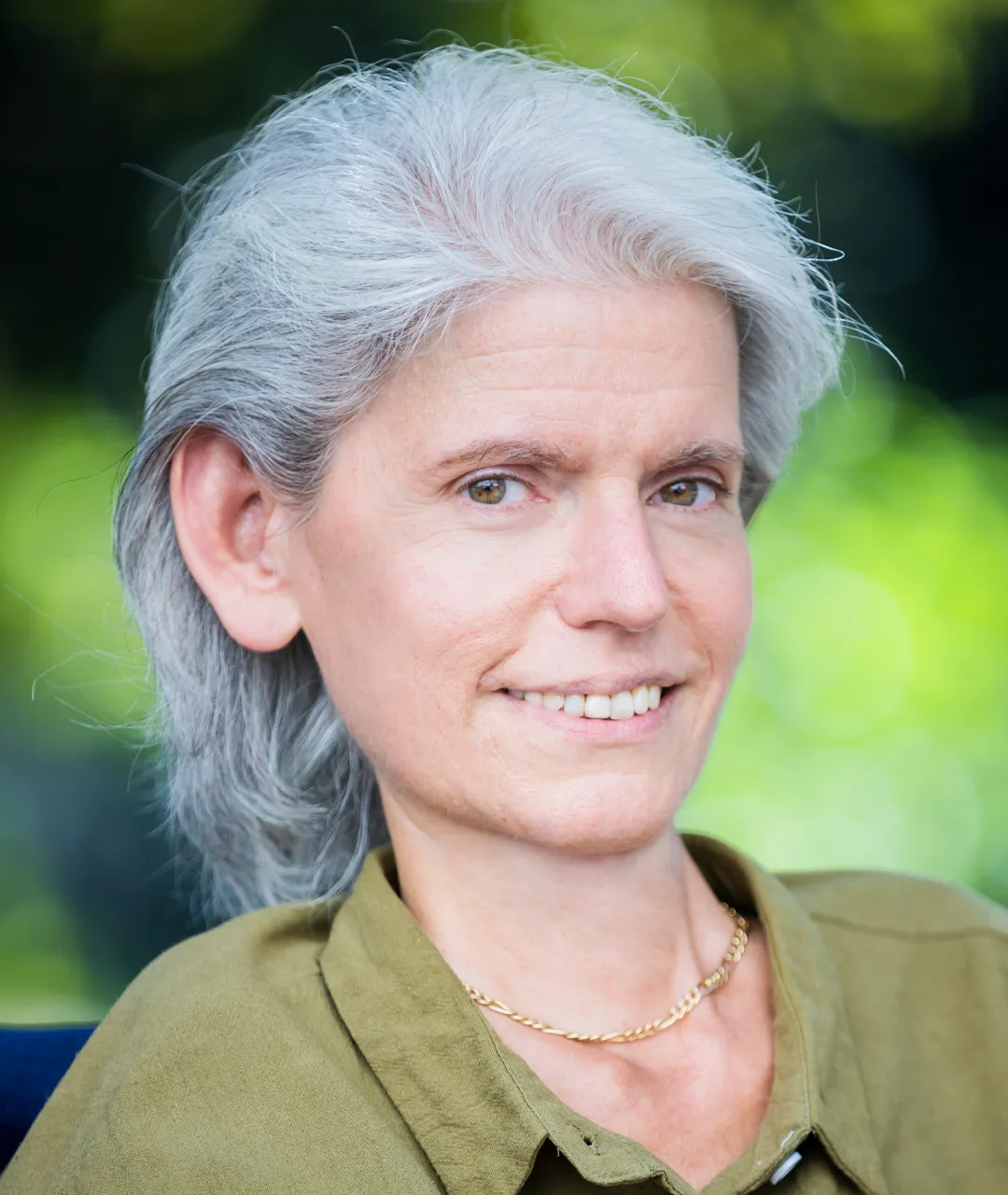 Frau Peric schaut seitlich in die Kamera und lächelt. Sie trägt schulterlanges, graues Haar, nach hinten gekämmt und eine olivfarbene Bluse. Sie hat eine Kette um den Hals und hellbraune Augen. Im Hintergrund sieht man unscharf grüne Pflanzen.
