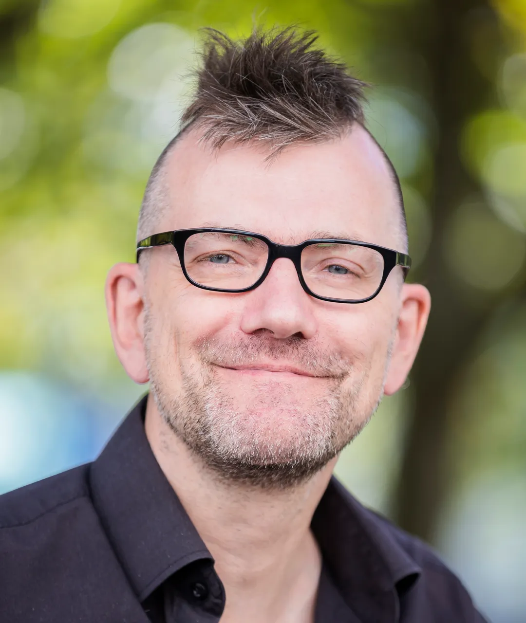 Herr Freels lächelt frontal in die Kamera. Er hat ein schwarzes Hemd an, trägt einen Dreitagebart und einen Irokesenschnitt. Sein Haar ist dunkelblond. Er hat eine schwarze Brille auf und blaue Augen. Im Hintergrund sieht man unscharf grüne Pflanzen.
