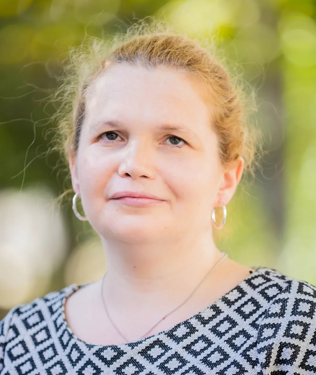 Frau Merslikin hat rotblondes Haar, das sie nach hinten gebunden trägt, und grün-blaue Augen. Sie trägt runde Kreolen in den Ohren.  Sie lächelt und trägt ein schwarz-weiß gemustertes Kleid und eine dünne, silberne Halskette. Im Hintergrund sieht man unscharf grüne Pflanzen.