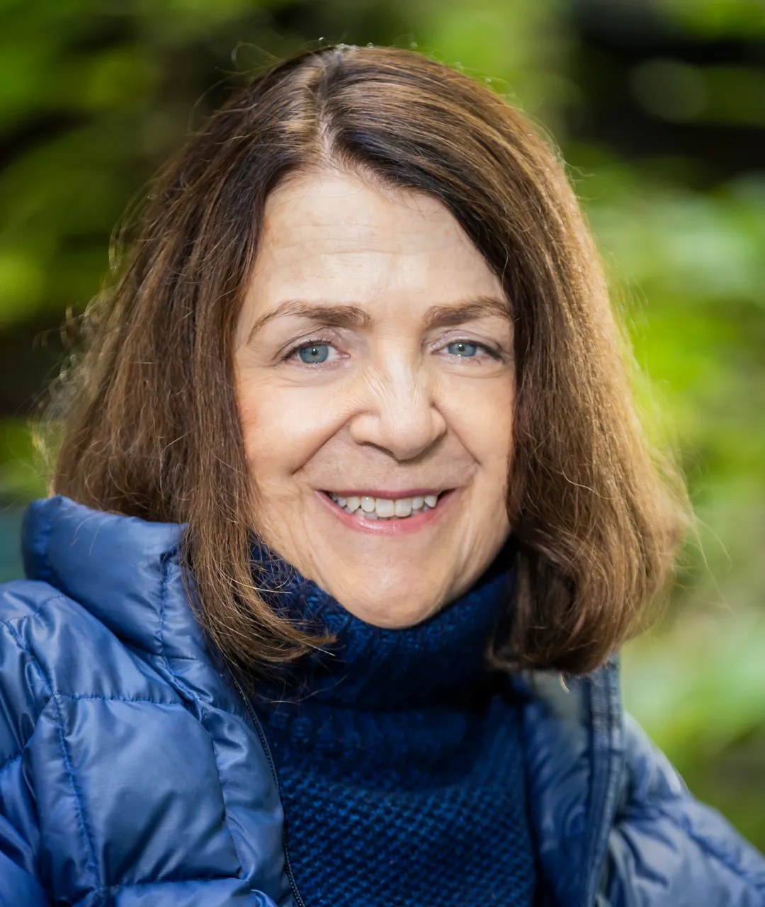 Frau Graudenz trägt ihr Haar offen. Sie hat schulterlanges, braunes Haar und blaue Augen. Sie hat einen dunkelblauen Wollpulli mit einem Rollkragen und eine offene, blaue Daunenjacke an. Im Hintergrund sieht man unscharf grüne Pflanzen.
