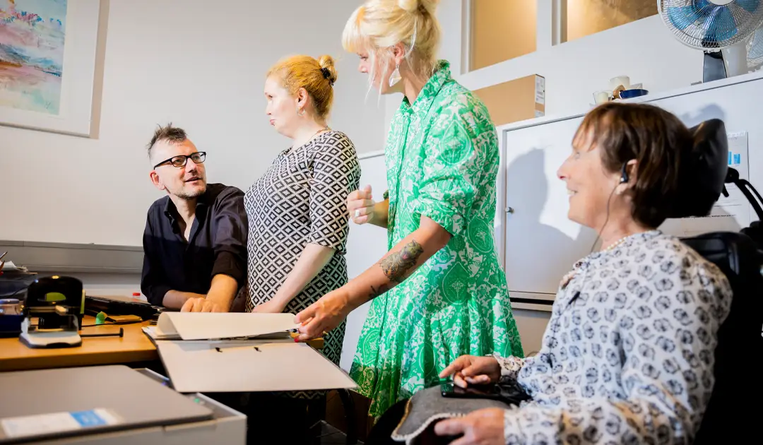 Besprechungssituation im Büro: rechts an der Wand sind Büroschränke, links im Bild ist der Schreibtisch. Zwischen Schreibtisch und Büroschrank sind vier Personen. Ganz links sitzt Herr Freels, daneben steht Frau Merslikin und ihre Assistentin, rechts im Vordergrund sitzt Frau Stenger. Auf dem Schreibtisch liegt ein aufgeschlagener Aktenordner, in dem die Assistenz blättert. Herr Freels schaut Frau Merslikin an und sagt etwas zu ihr. Frau Stenger sehen wir lachend im Vordergrund. Sie schaut die anderen Personen an.