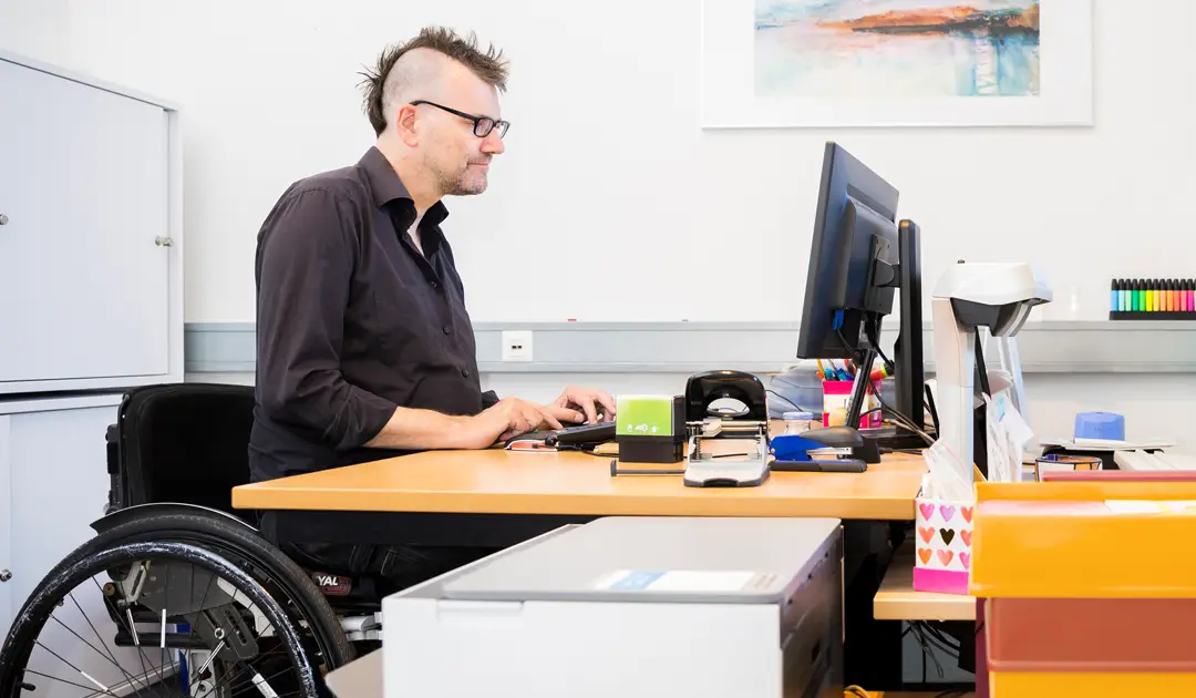 Bürosituation: Herr Freels sitzt am Schreibtisch und schreibt am Computer. Er trägt ein schwarzes Hemd.