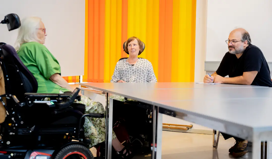Zu sehen ist eine Beratungssituation: im Hintergrund sieht man orangefarbene Lamellenvorhänge. Es gibt einen länglichen, grauen Tisch. Auf der linken Seite sitzt die Klientin, in der Bildmitte sitzt Frau Stenger, auf der rechten Seite vom Tisch sitzt Frau Stengers Assistenz.