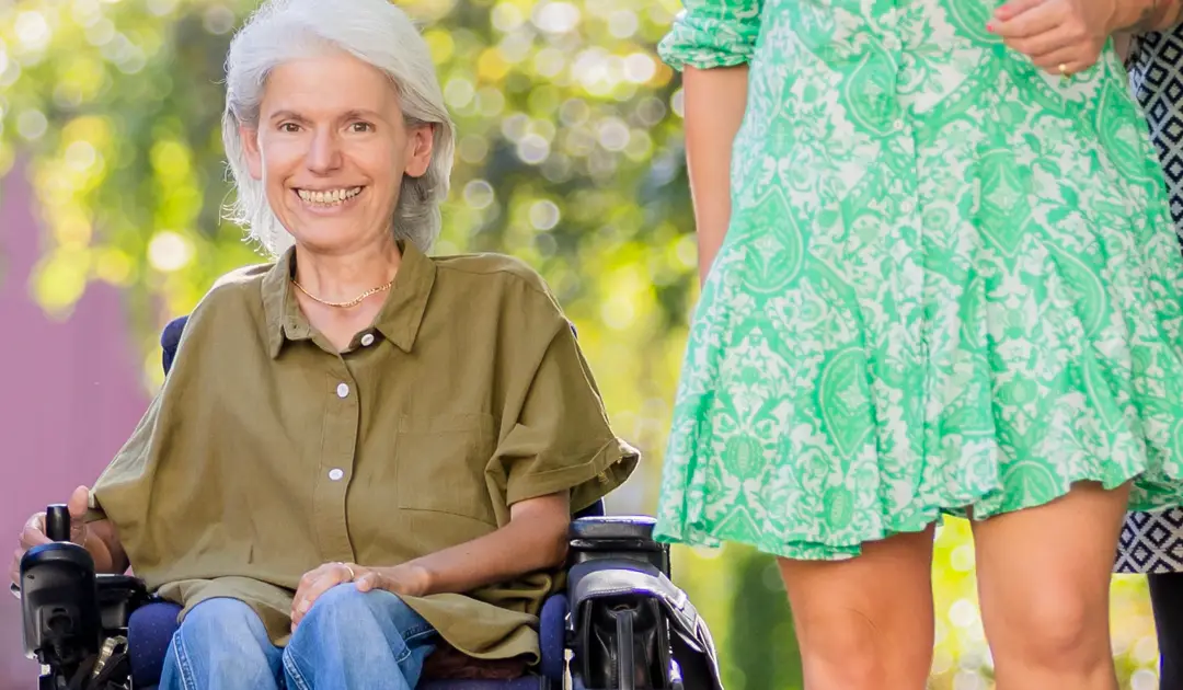 Auf der linken Bildhälfte sieht man Frau Peric in die Kamera lächeln. Sie trägt eine olivfarbene Bluse. Rechts von ihr im Anschnitt eine Assistentin, die ein hellgrünes Kleid trägt. Unscharf im Hintergrund sieht man Bäume.