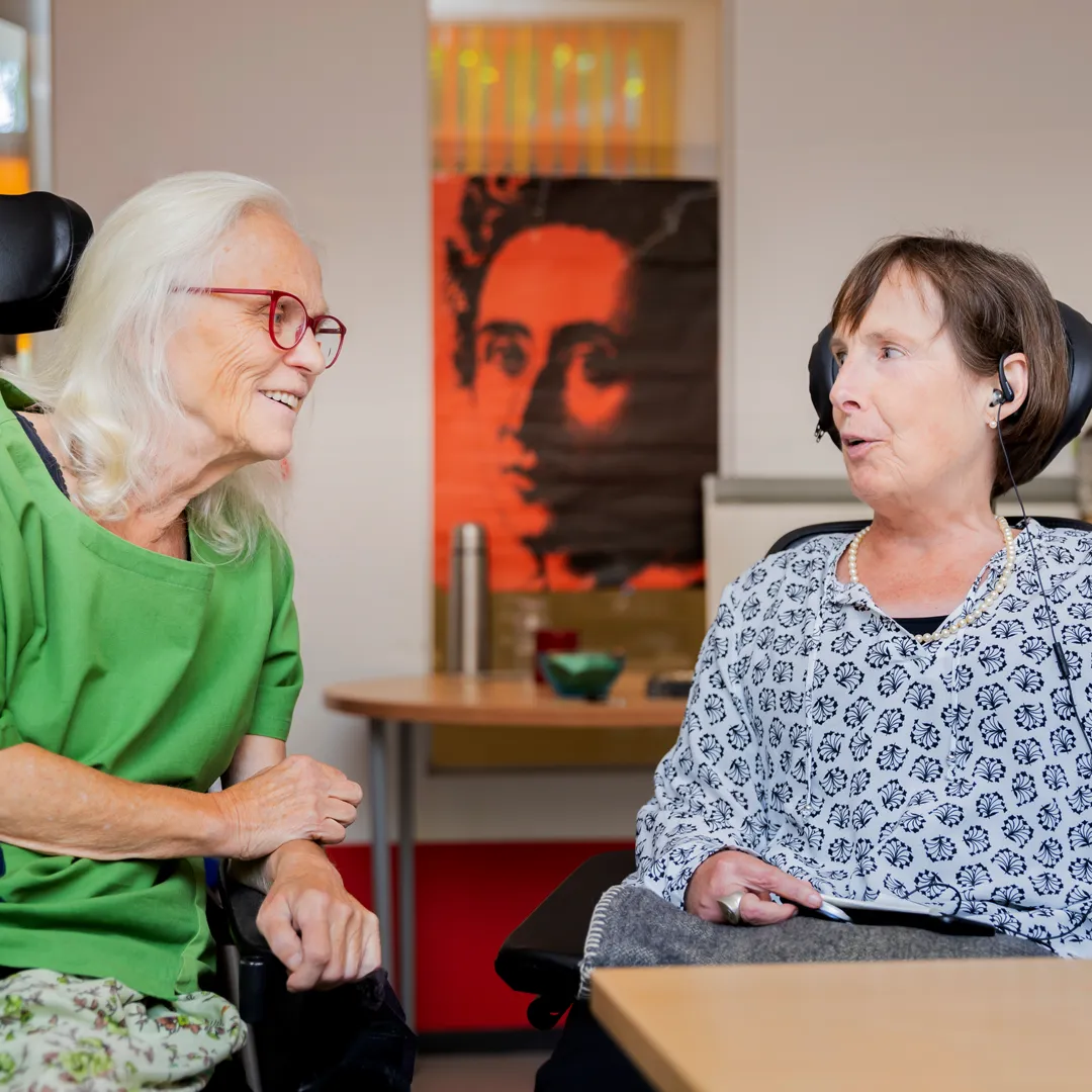 Bürosituation: Frau Stenger spricht mit einer Klientin. Unscharf im Hintergrund ist auf der rechten Seite ein Bücherregal zu sehen, zwischen den beiden Frauen hängt ein rotes Plakat mit Rosa-Luxemburg. Auf der linken Bildseite sitzt die Klientin, auf der rechten Seite sitzt Frau Stenger. Beide Frauen sitzen im Rollstuhl und sprechen angeregt miteinander.